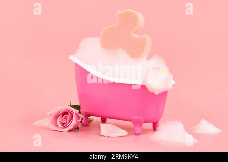 Petite baignoire avec mousse, fleur de rose et éponge sur fond rose Banque D'Images