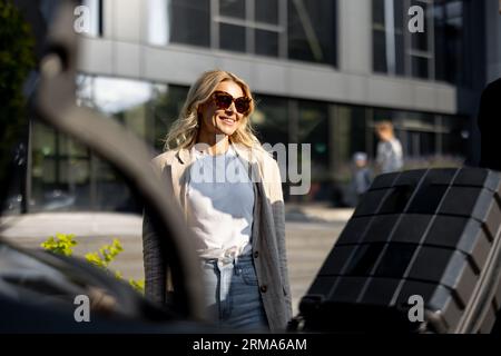 Femme d'affaires attend pendant que chauffeur emballe une valise dans le coffre de voiture Banque D'Images