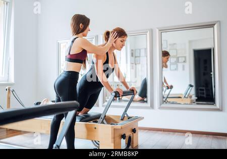 Jeune instructeur de pilates féminin aidant le client à ajuster la posture faisant des exercices de force de muscles abdominaux en utilisant la machine de reformer dans le gymnase athlétique de sport h Banque D'Images