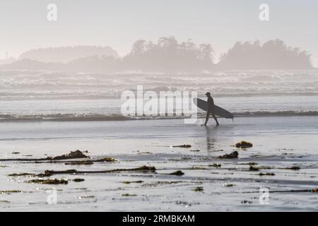 Silhouette de surfeur méconnaissable avec planche de surf marchant sur Cox Bay Beach près de l'océan Pacifique, Tofino, Île de Vancouver, Colombie-Britannique, Canada. Banque D'Images