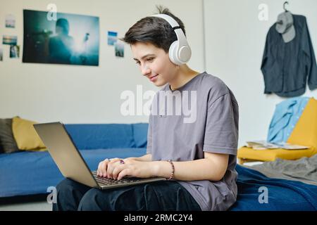 Brunette adolescente dans les écouteurs et les vêtements décontractés tapant sur le clavier d'ordinateur portable et regardant l'écran tout en étant assis sur le lit et le réseau Banque D'Images