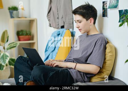 Joli adolescent en casualwear tapant sur le clavier d'ordinateur portable et regardant l'écran tout en faisant le plan d'un nouveau projet d'école dans l'environnement de la maison Banque D'Images