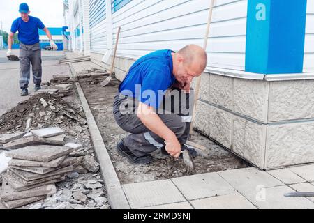Le maçon âgé pose des tuiles sur la fondation du bâtiment à l'extérieur. Entrepreneur en construction pose des tuiles de parement de façade de l'installation de production. L'homme pose des carreaux le jour d'été. Banque D'Images