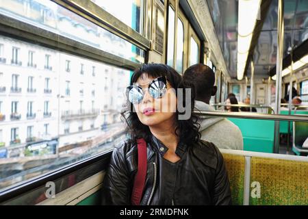 Une femme élégante dans une veste en cuir noir et des lunettes de soleil rondes miroir assise dans le train, une voiture sur une section surélevée du métro parisien Banque D'Images