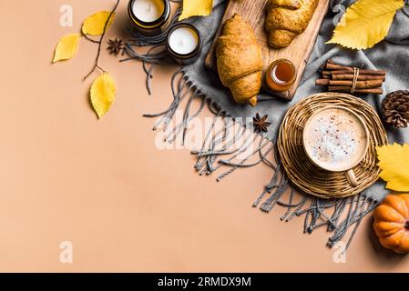 Concept esthétique d'automne. Tasse de café cappuccino avec croissants, bougies, plaid et feuilles jaunes d'automne sur fond beige, espace de copie. Banque D'Images