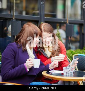 Deux filles dans un café en plein air buvant du café et utilisant un téléphone portable à Paris, France Banque D'Images