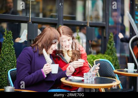 Deux filles gaies buvant du café, bavardant et utilisant leur téléphone portable dans un café de rue parisien Banque D'Images