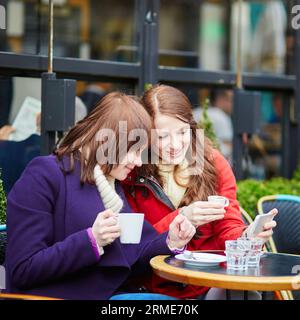 Deux filles dans un café en plein air buvant du café et utilisant un téléphone portable à Paris, France Banque D'Images