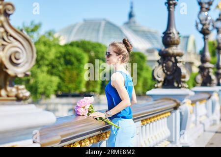 Belle jeune femme parisienne en blouse bleue avec bouquet de pivoines roses sur Pont Alexandre III à Paris, France Banque D'Images