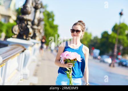 Belle jeune femme parisienne en blouse bleue avec bouquet de pivoines roses sur Pont Alexandre III à Paris, France Banque D'Images