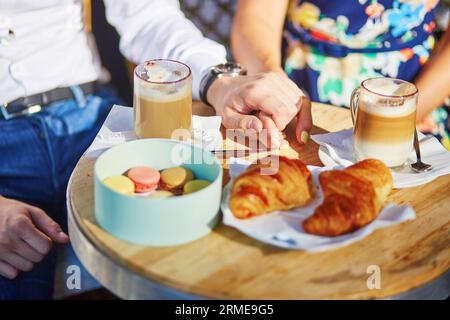 Gros plan de mains masculines et féminines lors d'un rendez-vous dans un café parisien. Couple tenant la main sur la table avec croissants, macarons et café Banque D'Images