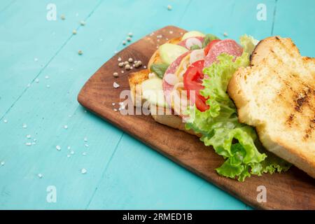 tranches de pain grillées avec concombre, oignon, tomates, laitue et salami, sandwich sur une planche à découper en bois, soft focus recadré vue latérale avec copie Banque D'Images