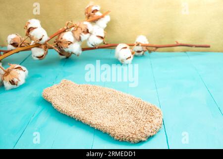 Toile de fond de produit de bain avec un gant de tissu de jute et des boutons de fleur de coton sur des planches de bois bleu et fond doré Banque D'Images