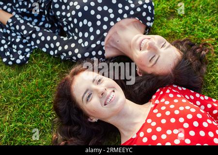 Belles sœurs jumelles en robes à pois rouges et noires allongées sur l'herbe près l'une de l'autre et souriantes Banque D'Images
