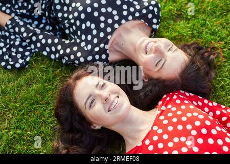 Belles sœurs jumelles en robes à pois rouges et noires allongées sur l'herbe près l'une de l'autre et souriantes Banque D'Images