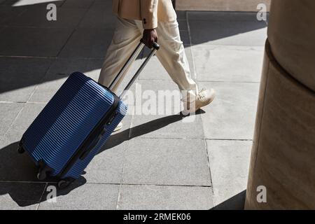 Photo recadrée d'un jeune homme d'affaires en pantalon blanc et baskets tirant une valise avec des bagages tout en se déplaçant le long des colonnes d'un bâtiment moderne Banque D'Images