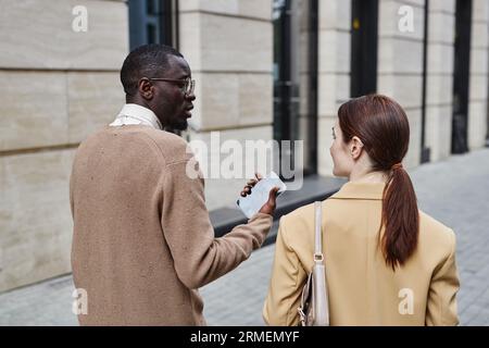 Vue arrière de deux jeunes employés interculturels en vêtements formalwear discutant tout en marchant le long d'un bâtiment moderne en milieu urbain Banque D'Images