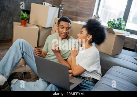 joyeuse femme afro-américaine parlant à son petit ami pendant les achats en ligne près des boîtes dans la nouvelle maison Banque D'Images