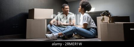 couple afro-américain avec du café à emporter parlant près des boîtes en carton dans la nouvelle maison, bannière Banque D'Images