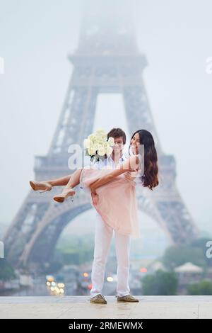 Beau couple romantique en amour avec bouquet de roses blanches s'amusant près de la tour Eiffel à Paris sur un jour de pluie nuageux et brumeux Banque D'Images