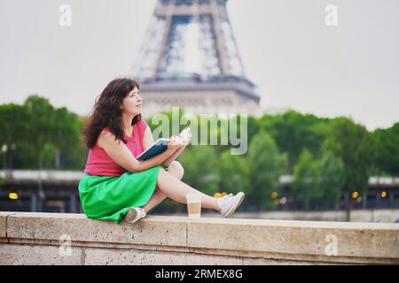 Jeune fille joyeuse avec du café pour aller lire un livre près de la tour Eiffel. Etudiant se préparant à l'examen à Paris. Prog. D'échange d'éducation internationale Banque D'Images