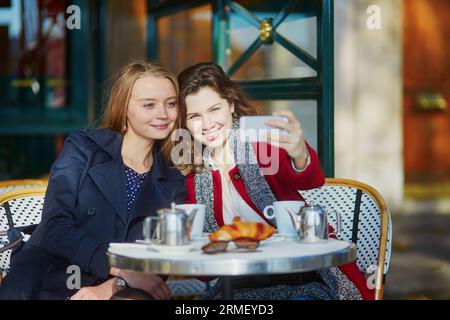 Deux jeunes filles dans un café en plein air parisien, buvant un café avec un croissant et prenant un selfie avec un téléphone portable. Concept d'amitié Banque D'Images