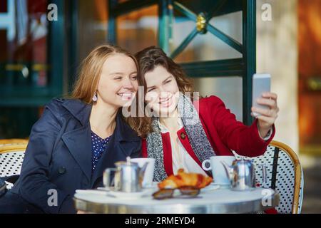 Deux jeunes filles dans un café en plein air parisien, buvant un café avec un croissant et prenant un selfie avec un téléphone portable. Concept d'amitié Banque D'Images