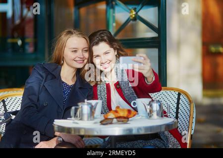 Deux jeunes filles dans un café en plein air parisien, buvant un café avec un croissant et prenant un selfie avec un téléphone portable. Concept d'amitié Banque D'Images