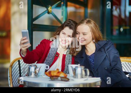 Deux jeunes filles dans un café en plein air parisien, buvant un café avec un croissant et prenant un selfie avec un téléphone portable. Concept d'amitié Banque D'Images