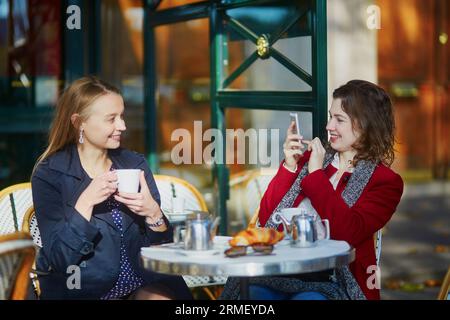 Deux jeunes filles dans un café en plein air parisien, buvant un café avec un croissant et prenant un selfie avec un téléphone portable. Concept d'amitié Banque D'Images