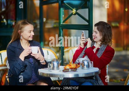 Deux filles dans un café en plein air buvant du café et utilisant un téléphone portable à Paris, France Banque D'Images