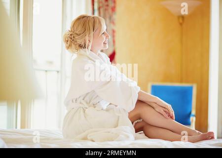 Belle jeune femme en peignoir blanc assise sur le lit tôt le matin et s'étirant après le sommeil de nuit Banque D'Images