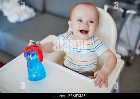 Petite fille de bébé assise dans une chaise haute à la maison ou au restaurant et boire de l'eau de tasse sippy Banque D'Images