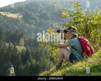 Touriste féminin assis sur la colline, admirant la vue sur la montagne à travers des jumelles Banque D'Images