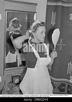 Dans les années 1940 Une jeune femme travaillant comme femme de ménage se regarde dans le miroir et met ses cheveux propres et rangés. Elle est soigneusement vêtue d'un tablier blanc. Suède 1940. Kristoffersson ref 56-6 Banque D'Images