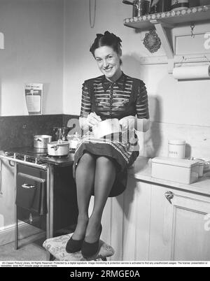 Dans la cuisine 1940s. Intérieur d'une cuisine et une jeune femme sourit à la caméra assise avec une casserole dans le genou et fouette le contenu. Suède 1945 Kristoffersson ref N138-3 Banque D'Images