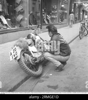 In the 1950s. A young man with his Triumph motorcycle that for some reason has some engine troubles and he is seen fixing something with it in the street. He is tough looking wearing a typical leather jacket with padded shoulders and the logotype of Triumph written on it. Sweden 1958 Kristoffersson ref CA29-12 Stock Photo