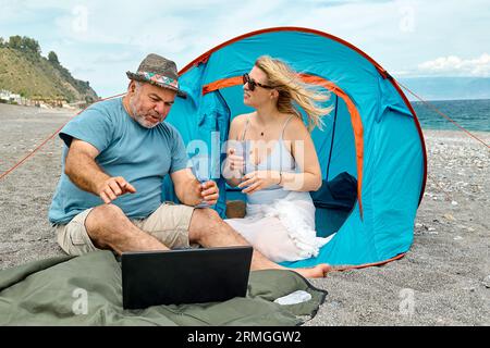 Couple s'amusant, boire du vin et parler avec des amis dans l'appel vidéo dans l'ordinateur portable en pique-nique près de tente sur le bord de la mer. Vacances d'été sur la plage à outd Banque D'Images