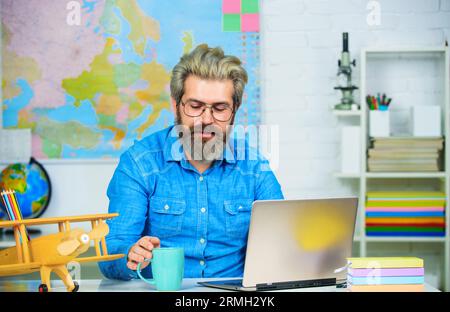 Professeur souriant buvant du café en classe. L'homme barbu boit du café dans la salle des professeurs. Professeur d'université travaillant avec un ordinateur portable ont l'école Banque D'Images