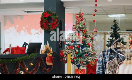 Sapin de Noël joliment décoré dans le magasin de vêtements prêt à répandre la joie de vacances aux clients visitant qui cherchent à acheter des cadeaux. Noël ornent la boutique de mode dans le centre commercial pendant la saison des vacances d'hiver Banque D'Images