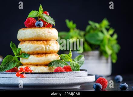 Crêpes épaisses au fromage cottage aux myrtilles, framboises et groseilles rouges, décorées de menthe fraîche sur une assiette grise, délicieux petit déjeuner. Tabl. Noir Banque D'Images