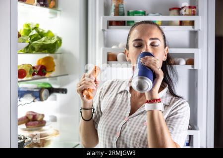 Femme affamée en pyjama boit de la bière et mange des saucisses par le réfrigérateur la nuit. Banque D'Images