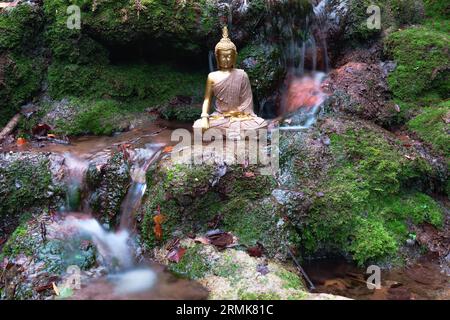 Statue de Bouddha assis dans une rivière avec des rochers et de la mousse verte et avec une cascade avec effet d'eau éclaboussant Banque D'Images