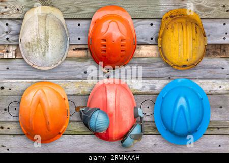 Six casques de construction vieux et usés colorés accrochés à un mur en bois Banque D'Images