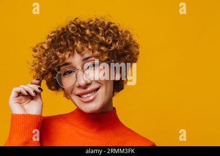 Joyeuse femme de gingembre d'âge moyen dans des lunettes touchant les cheveux isolés sur fond de studio jaune Banque D'Images