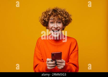 Surprise joyeuse femme de gingembre d'âge moyen utilisant smartphone isolé sur fond jaune Banque D'Images