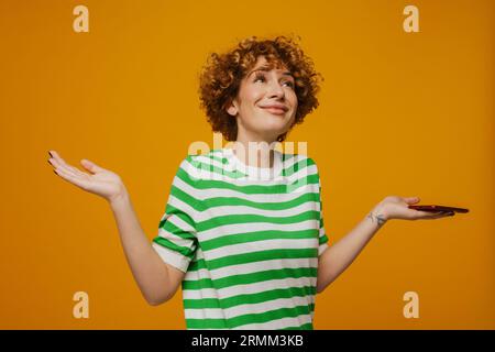 Femme bouclé de gingembre dans un t-shirt rayé gestant et utilisant un téléphone portable isolé sur fond jaune Banque D'Images