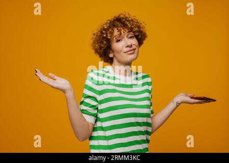 Femme bouclé de gingembre dans un t-shirt rayé gestant et utilisant un téléphone portable isolé sur fond jaune Banque D'Images