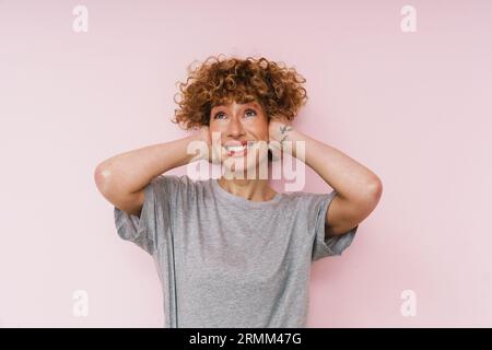 Gingembre frisé femme posant avec sourire et couvrant ses oreilles isolé sur fond rose Banque D'Images