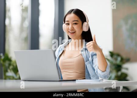 Heureuse étudiante asiatique recommandant un cours d'étude en ligne, montrant le pouce vers le haut et souriant à la caméra Banque D'Images